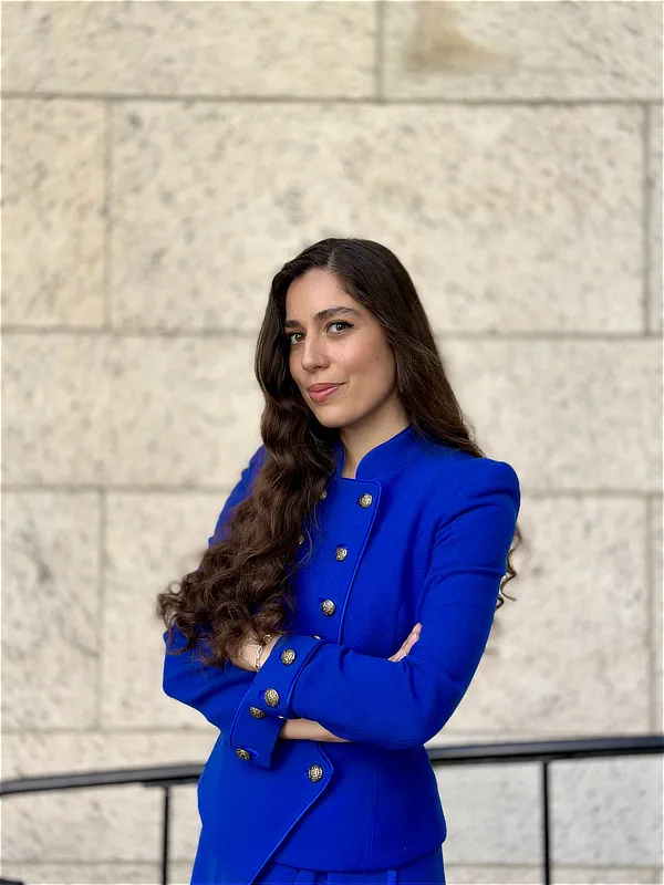 Retrato profesional en exterior de una mujer con chaqueta azul cruzada, brazos cruzados y expresión confiada, posando ante una pared de piedra clara.