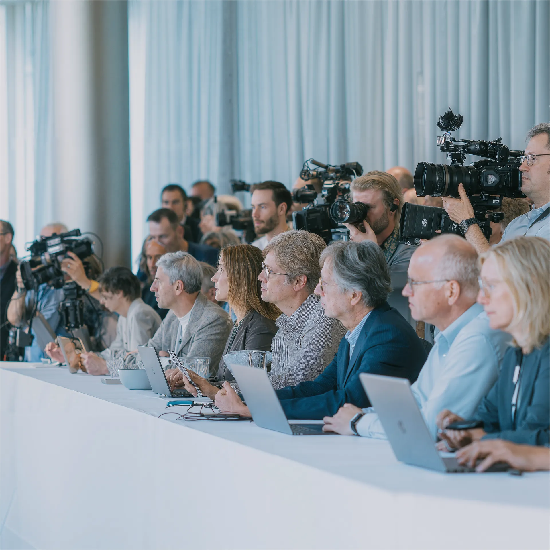 Periodistas y fotógrafos en una rueda de prensa, tomando notas y grabando con cámaras profesionales desde una mesa cubierta de ordenadores portátiles.