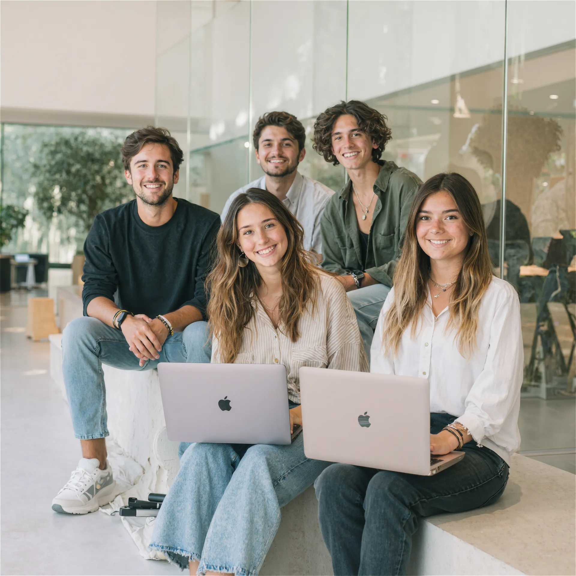 Cinco jóvenes en un espacio acristalado y luminoso, sentados en bancos claros y usando portátiles mientras miran a cámara.
