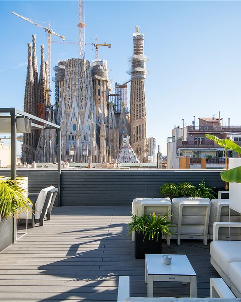 Terraza moderna con muebles exteriores y plantas, situada frente a la Sagrada Familia, ofreciendo vistas privilegiadas del emblemático monumento y del skyline urbano de Barcelona.