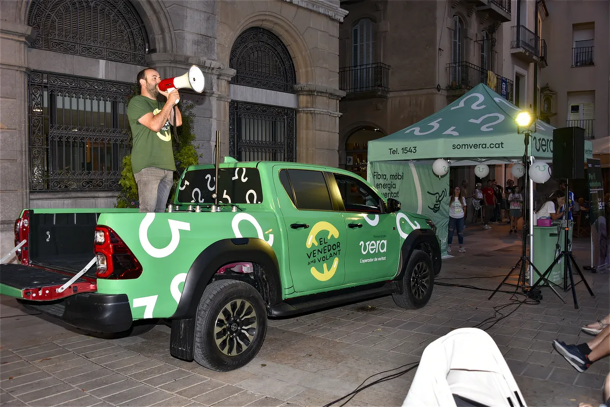 Evento en la calle con vehículo corporativo de Vera y un presentador usando megáfono sobre la camioneta, interactuando con asistentes cerca de una carpa promocional.