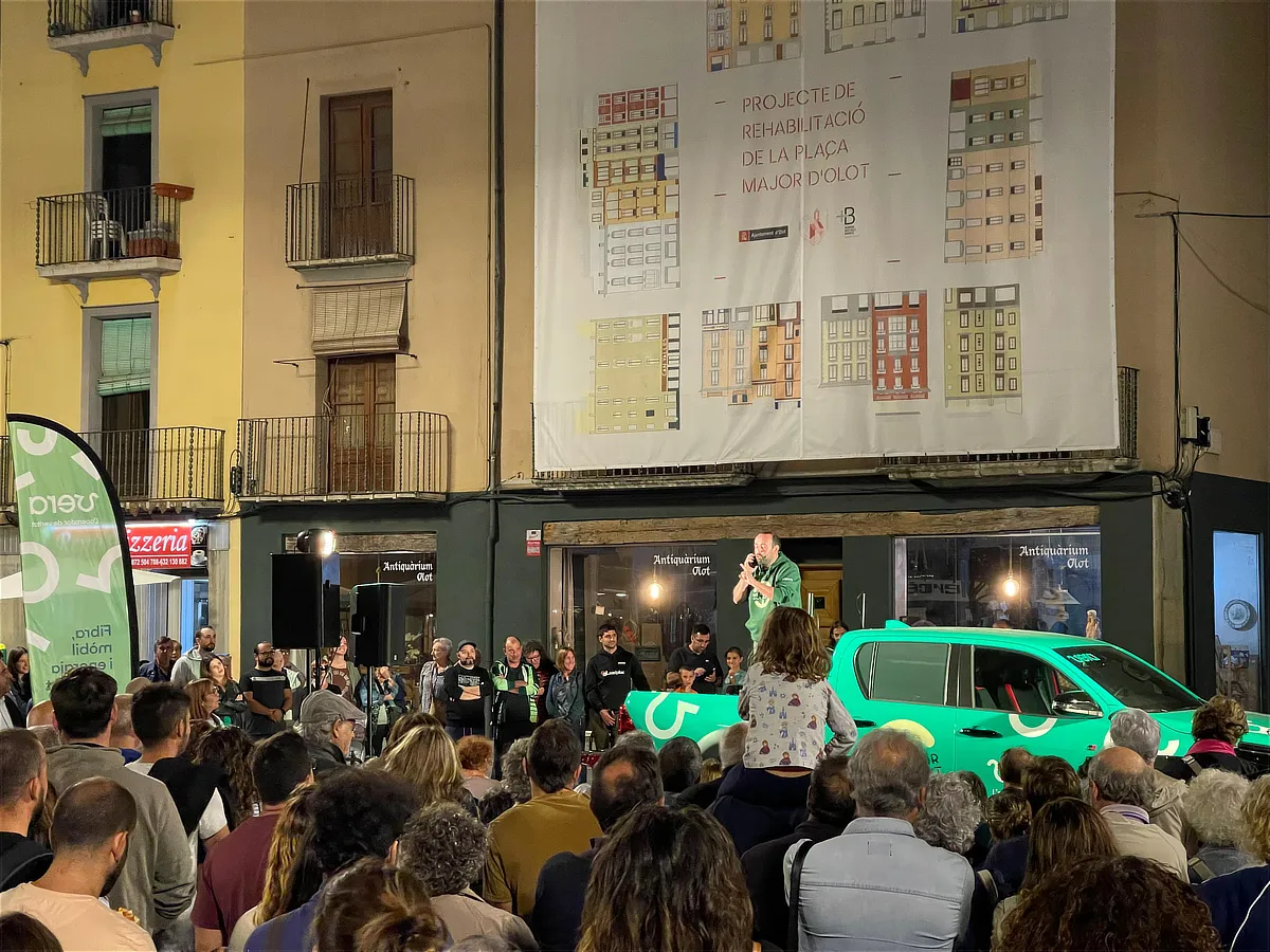 Acto público en una plaza con numeroso público asistiendo mientras un presentador habla desde la caja de una camioneta verde de Vera, rodeado de comercios y edificios históricos.