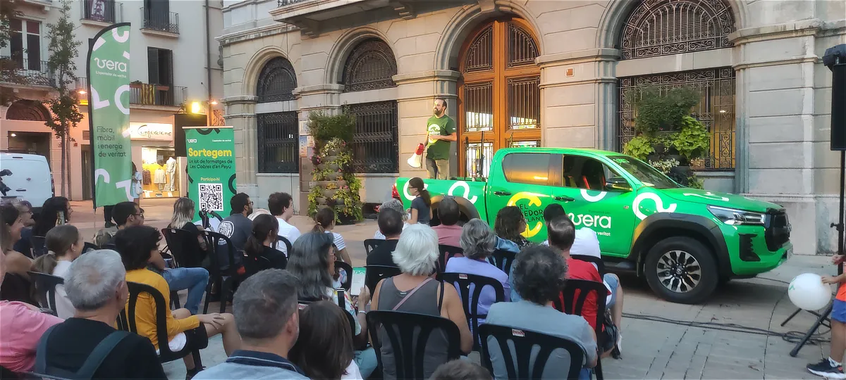 Presentación al aire libre con público sentado frente a un portavoz de Vera que usa megáfono sobre una camioneta corporativa, junto a cartelería promocional y banderolas de marca.