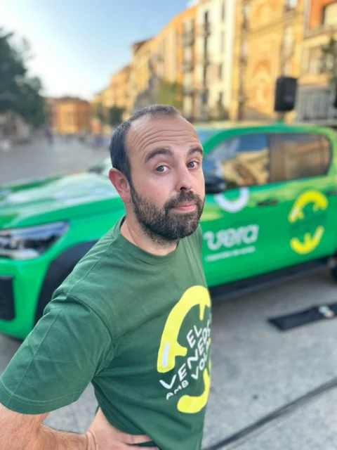 Peyu posando en primer plano con camiseta verde frente a un vehículo corporativo también verde, en una calle urbana con edificios y luz natural al fondo.